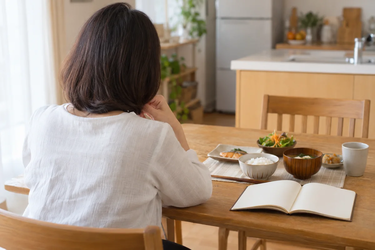 食卓で食事とノートを前に考える女性の後ろ姿。イメージ写真
